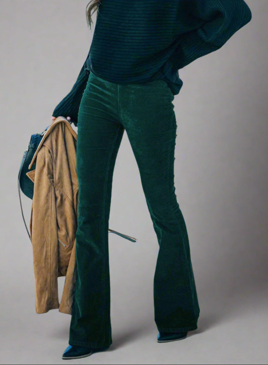Vrouw draagt donkergroene flared broek met zwarte trui, houdt bruine jas vast. Modieuze herfstoutfit, trendy dameskleding, straatstijl.