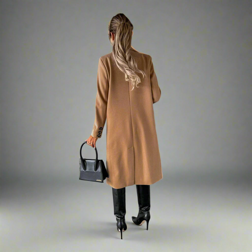 Vrouw in stijlvolle camel winterjas, zwarte laarzen en handtas, staand tegen een betonnen muur. Wintermode, dameskleding, trendy outfit.