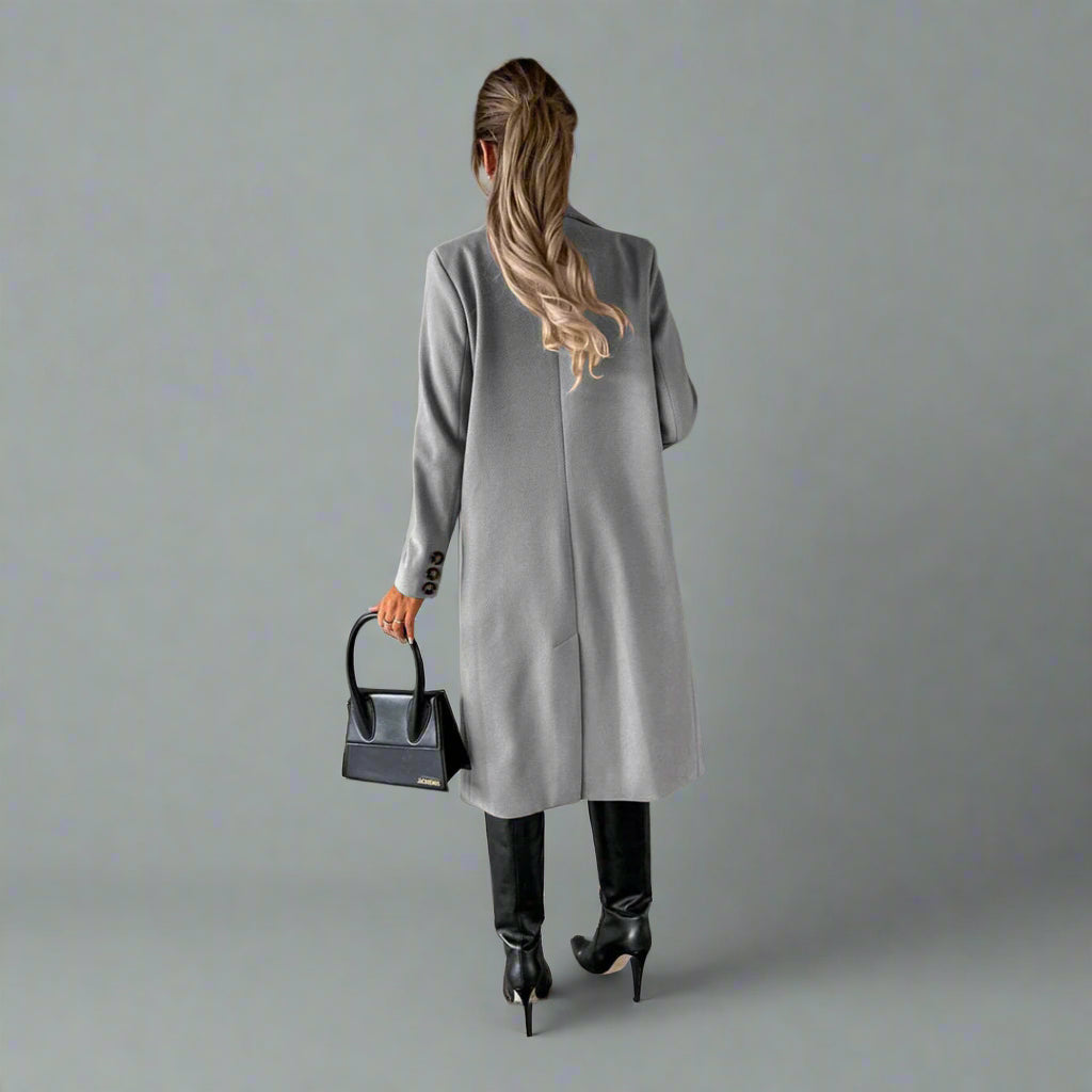 Vrouw in grijze winterjas, zwarte leren laarzen en handtas, staand tegen betonnen muur. Stijlvolle wintermode, minimalistisch design.