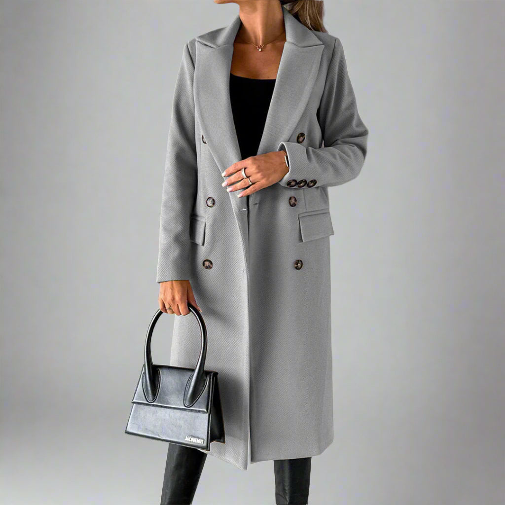 Grijze dames winterjas met dubbele knopenrij, gedragen met zwarte handtas en zwarte broek. Stijlvolle wintermode, elegante jas voor dames.