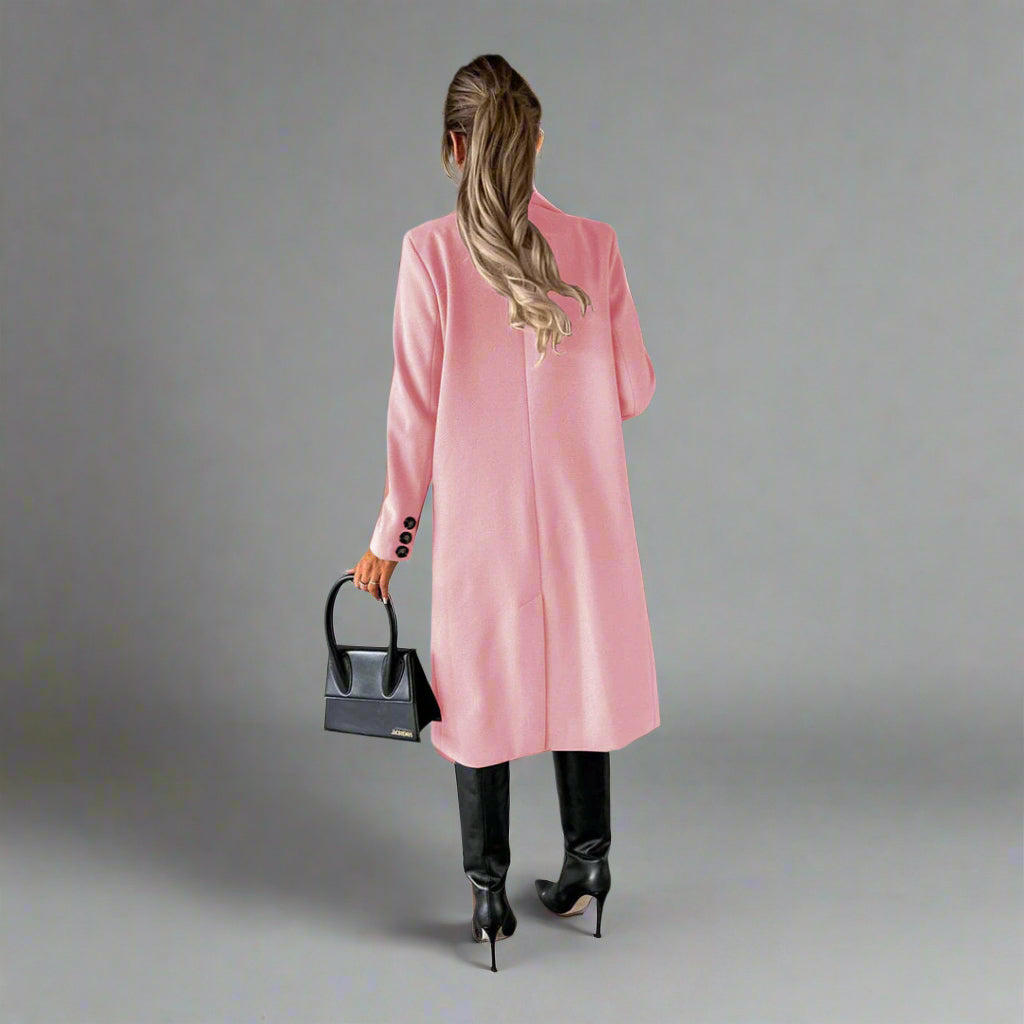 Vrouw in roze winterjas met zwarte laarzen en handtas, staand tegen een betonnen muur. Stijlvolle damesmode, winterkleding, trendy outfit.