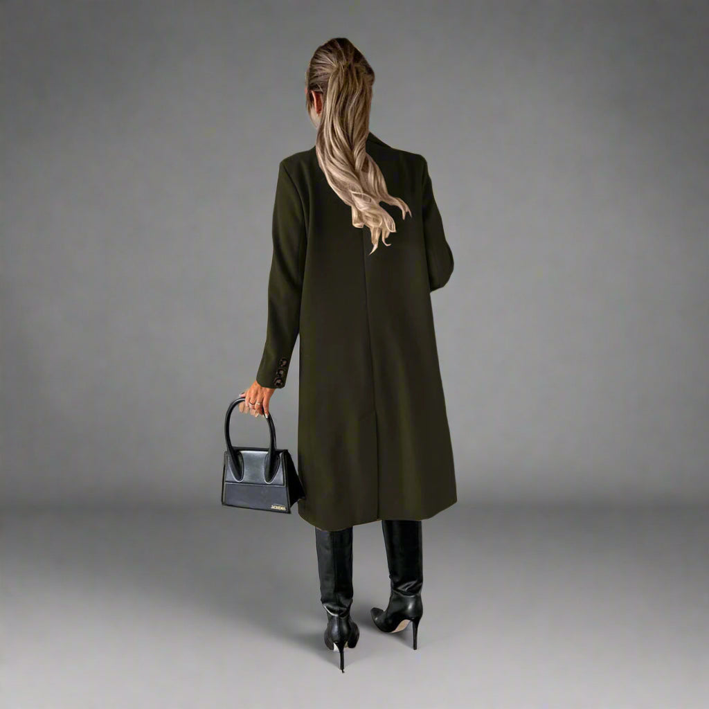 Vrouw in groene winterjas met zwarte laarzen en handtas, staand tegen een betonnen muur. Stijlvolle wintermode, trendy dameskleding.