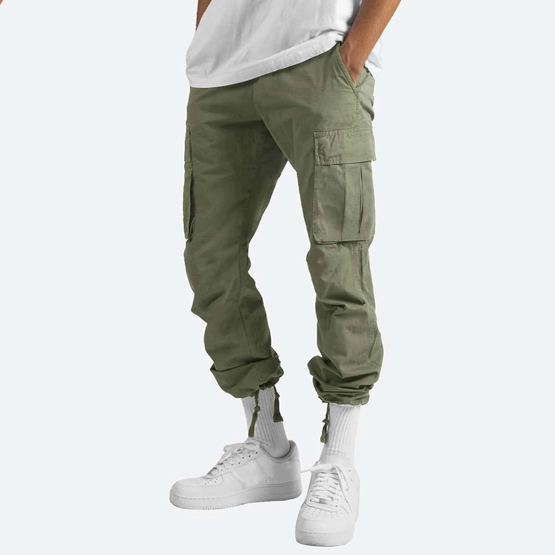 Olijfgroene cargobroek voor heren met meerdere zakken, casual stijl, gecombineerd met witte sneakers en sokken. Perfect voor streetwear en outdoor activiteiten.