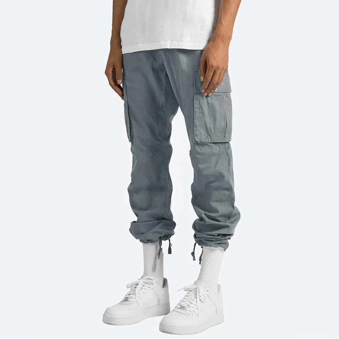 Grijze cargobroek voor heren met zakken, casual streetwear stijl, gecombineerd met witte sneakers en sokken. Perfect voor modieuze, comfortabele outfits.