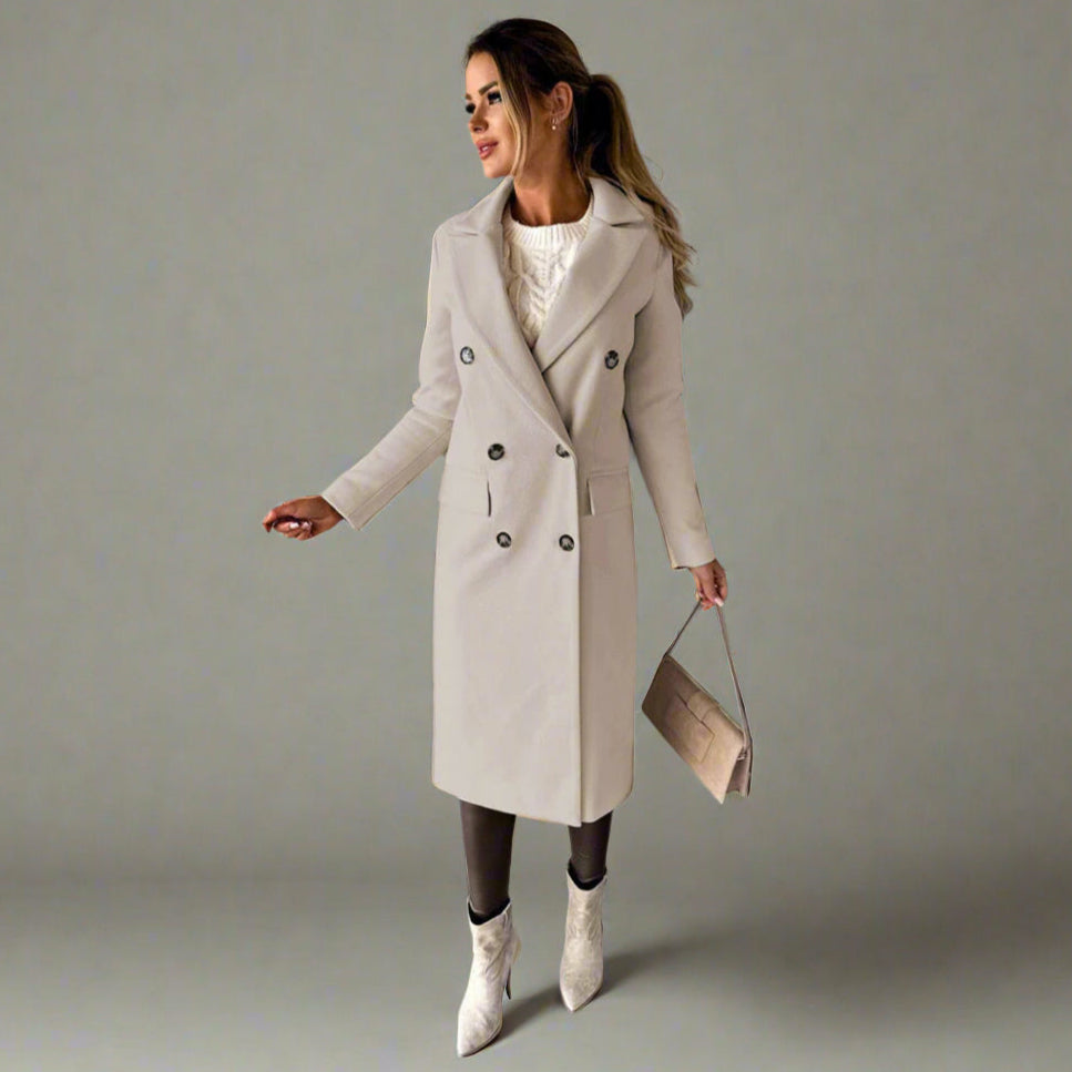 Vrouw in stijlvolle beige winterjas met dubbele rij knopen, witte trui en laarzen, poseert tegen een betonnen muur. Wintermode, dameskleding.